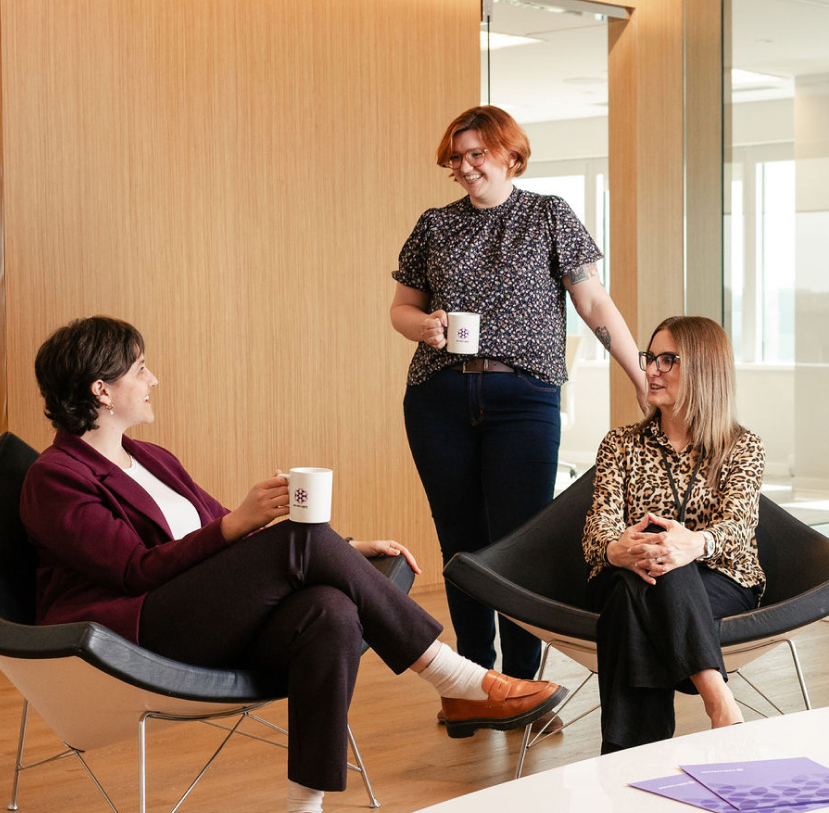 Three team members share a casual conversation in an office lounge, holding BOYNECLARKE mugs.